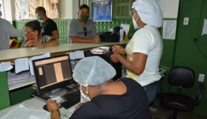 Enfermeira mede a altura de uma criança em unidade de saúde enquanto mãe observa.