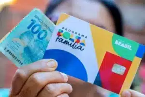 Mão com cartão Bolsa Família e celular com app Caixa Tem.