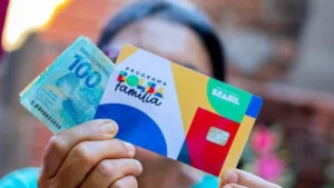 Mão segurando celular com o aplicativo Caixa Tem aberto, indicando recebimento de Bolsa Família e Pé-de-Meia