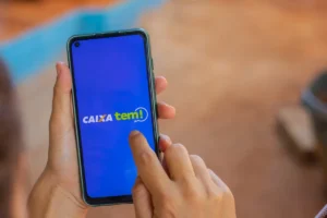 Estudante do ensino médio sorri enquanto confere o aplicativo Caixa Tem em seu celular, com uma sala de aula ao fundo.