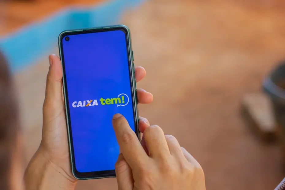 Estudante do ensino médio sorri enquanto confere o aplicativo Caixa Tem em seu celular, com uma sala de aula ao fundo.