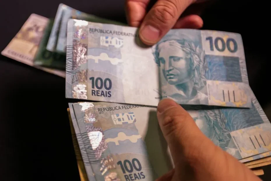 Mãos de trabalhador brasileiro consultando o aplicativo Carteira de Trabalho Digital para verificar informações do PIS/Pasep, com notas de real ao fundo.