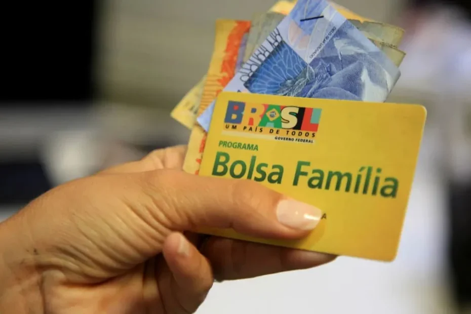 Cartão do Bolsa Família sendo segurado por uma beneficiária em frente a uma unidade da Caixa.