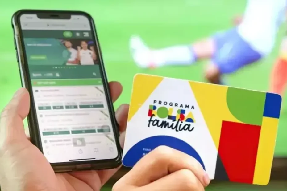 Mãos segurando celular com tela de aposta online e símbolo de bloqueio, representando a proibição para beneficiários de programas sociais.