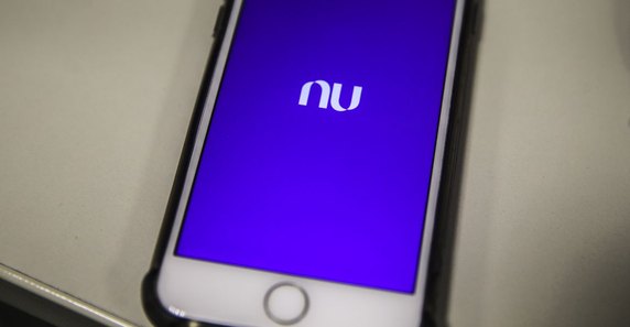 Pessoa olhando o aplicativo do Nubank no celular, com o logo da HBO Max ao fundo, simbolizando a mudança nos benefícios.