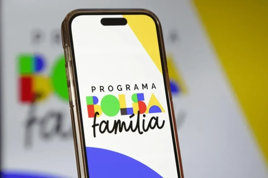 Mão usando o aplicativo Caixa Tem para consultar o Bolsa Família, com o cartão do benefício ao lado e uma família ao fundo.