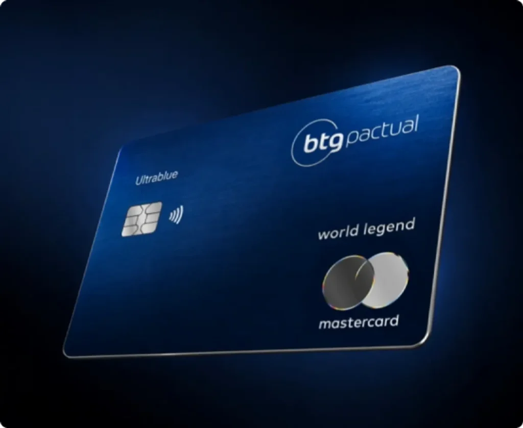 Mão elegante segurando o cartão de crédito BTG Pactual Ultrablue World Legend em um lounge VIP de aeroporto, com taça de vinho e ambiente luxuoso ao fundo.