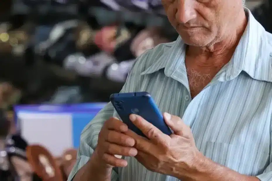 Mãos segurando um celular com tela de aplicativo de apostas bloqueado, com um cartão de benefício social ao fundo, simbolizando proteção financeira.