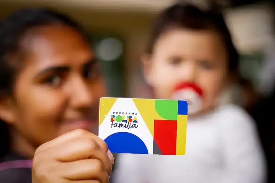 Cartão do Bolsa Família sendo segurado em frente a uma agência da Caixa.